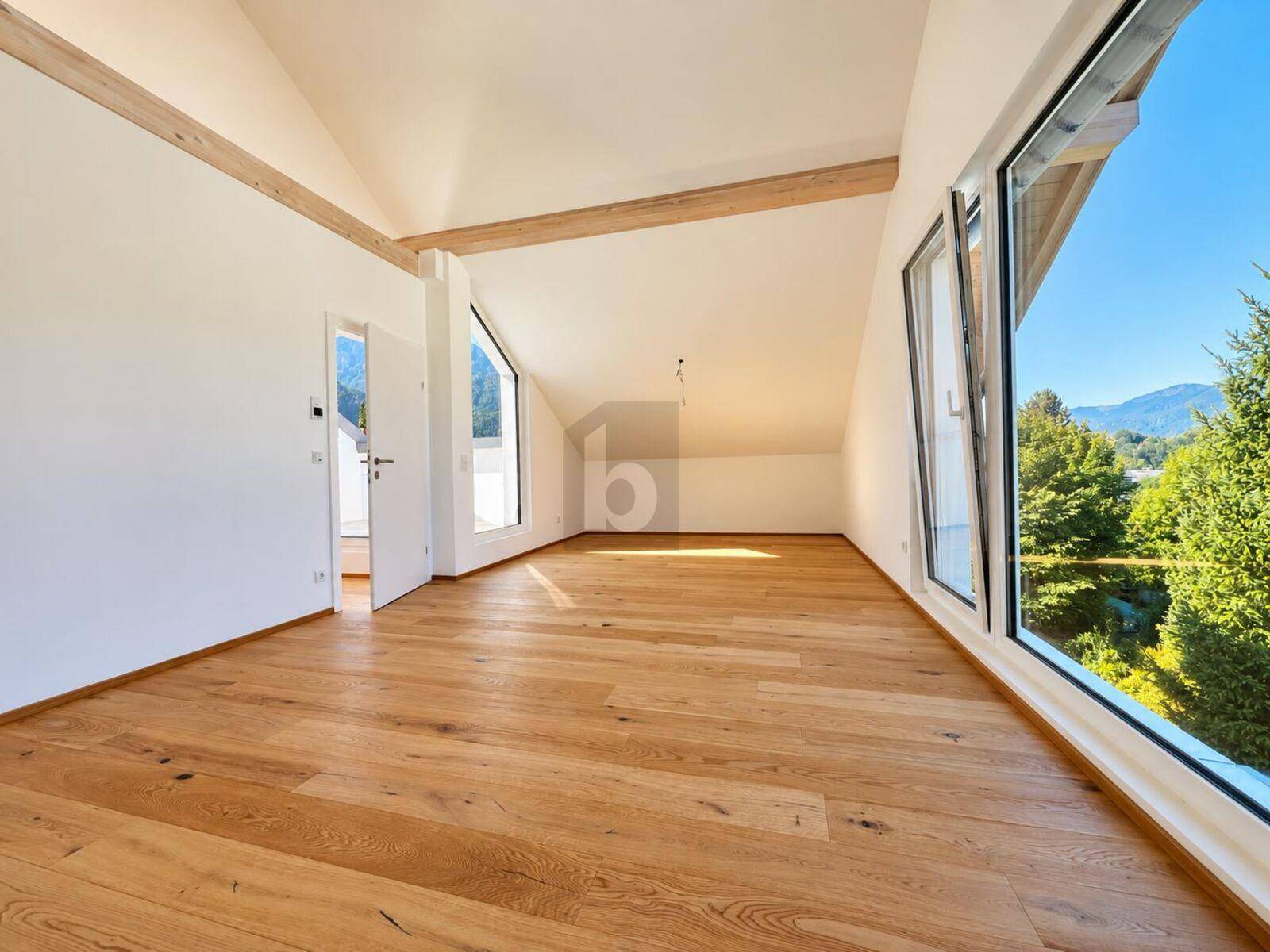 Helles Zimmer mit Holzboden, schrägen Decken und großen Fenstern mit Blick ins Grüne und auf Berge.