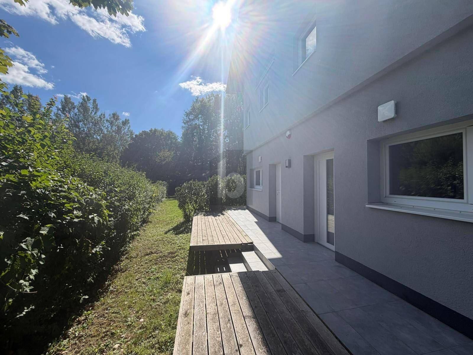 Sonnige Terrasse mit Holzboden und Fliesenbereich, umgeben von grünen Hecken und Bäumen.