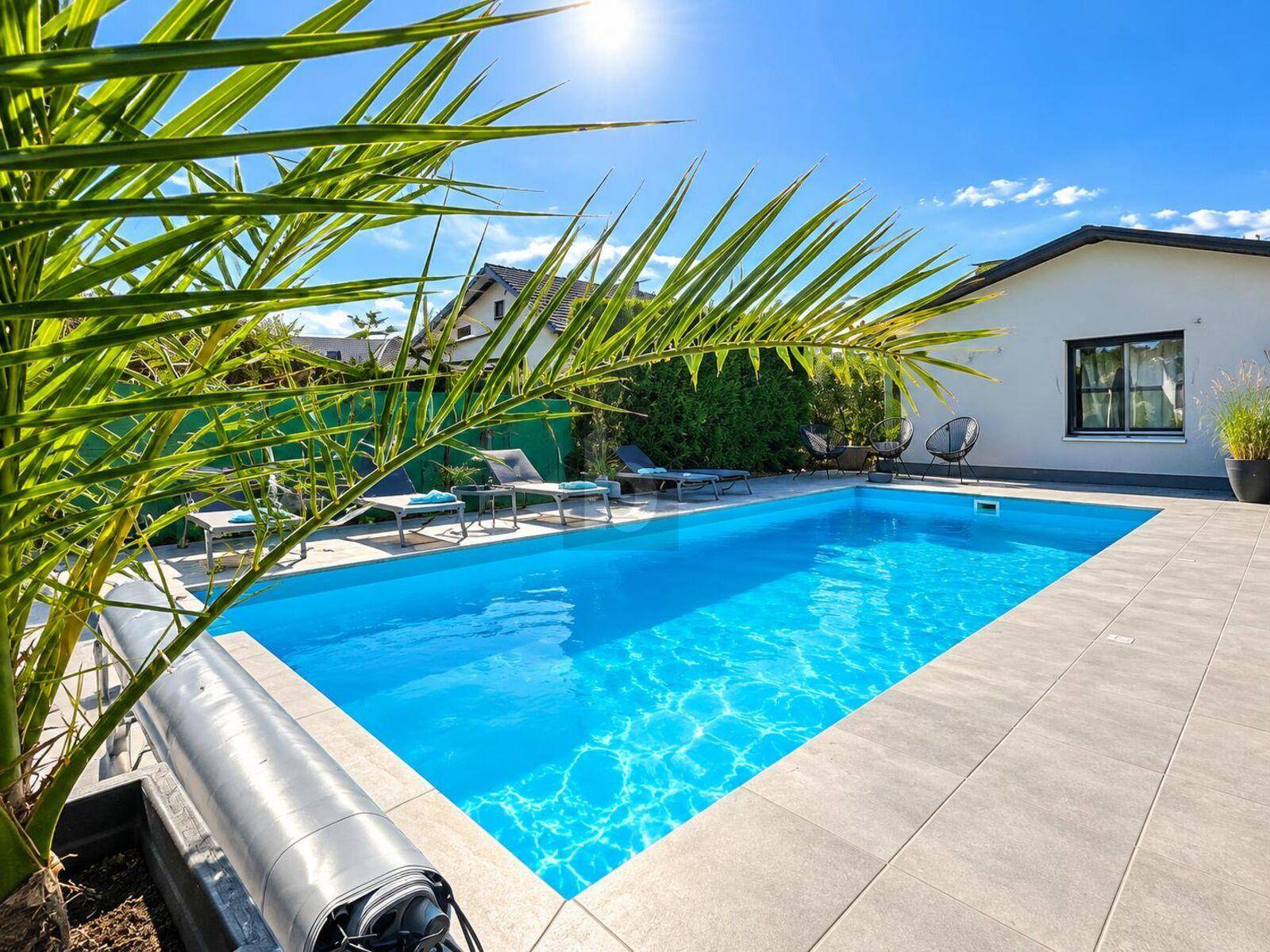 Sonniger Gartenbereich mit großem Swimmingpool, Liegestühlen und Palmen.