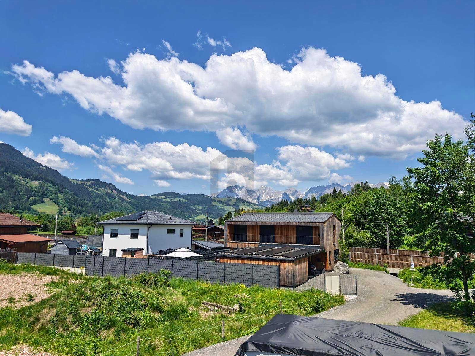 Panoramablick auf Wohnhäuser in einer grünen Landschaft mit majestätischen Bergen im Hintergrund unter blauem Himmel.