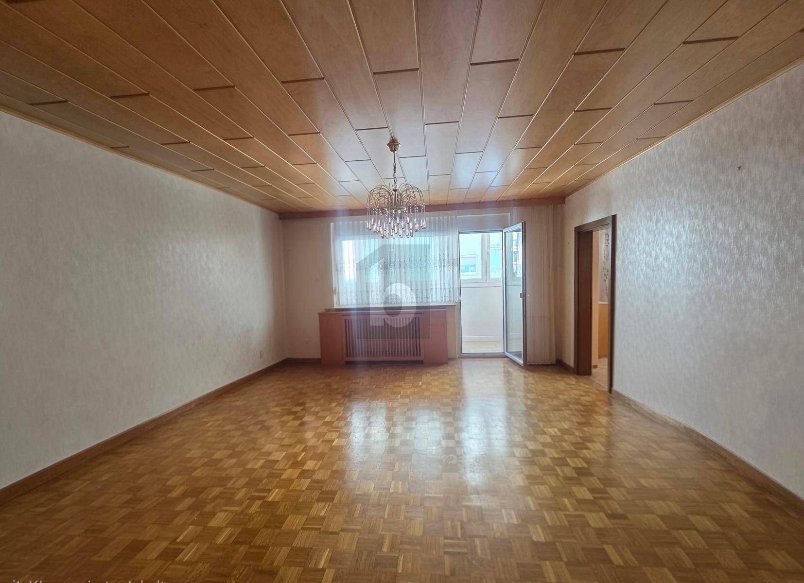 Geräumiges Wohnzimmer mit Parkettboden, Holzdecke und Zugang zu einem Balkon.