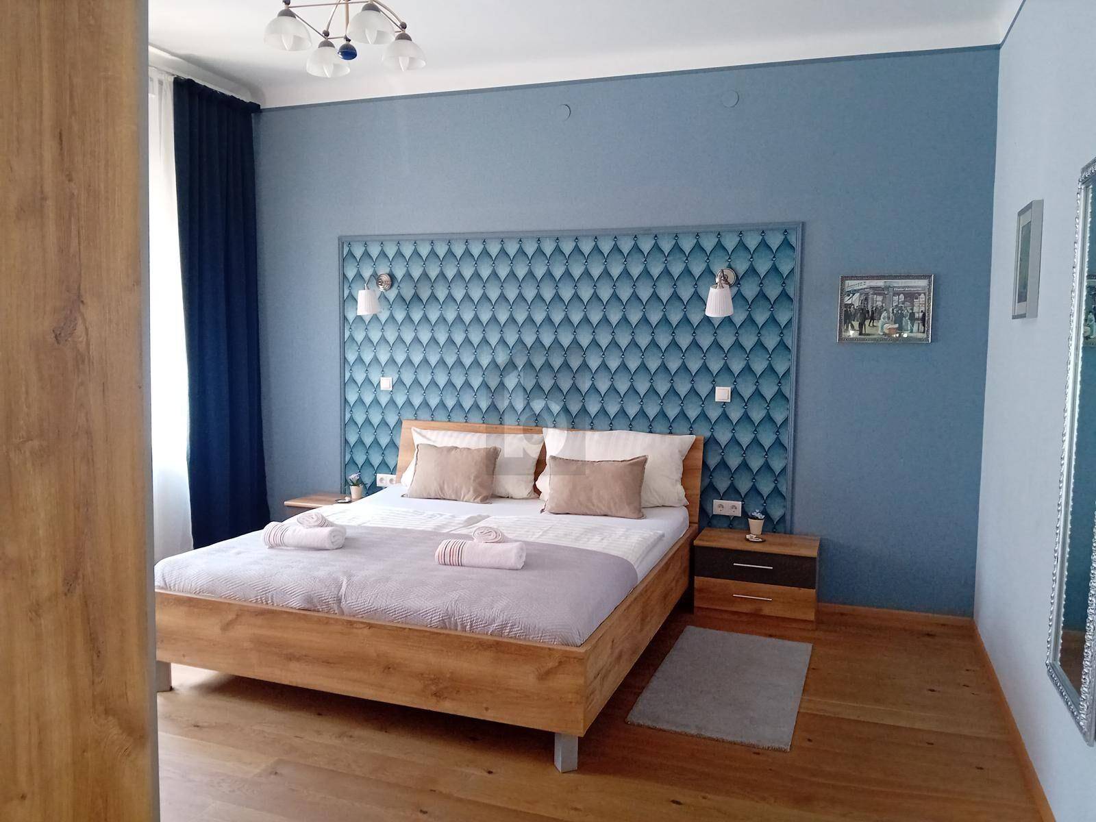 Modernes Schlafzimmer mit blauer Akzentwand, Holzbett und hellem Parkettboden, gut beleuchtet durch ein Fenster.