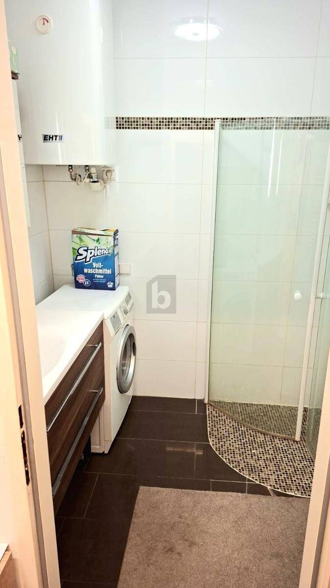 Funktionales Badezimmer mit Dusche, Waschbecken, Waschmaschine und weißen Fliesen an Wänden und Boden.