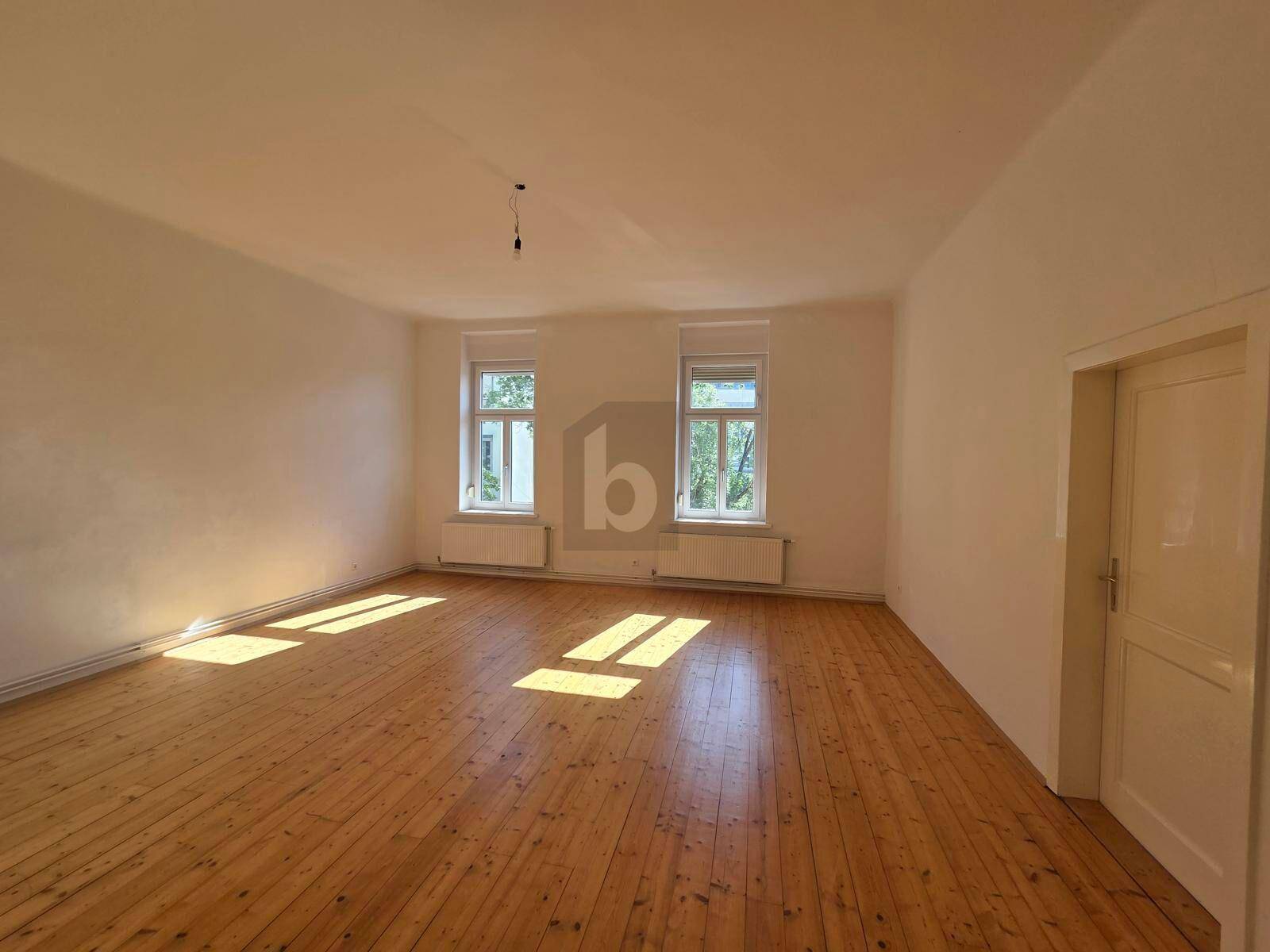 Heller, leerer Raum mit Holzdielenboden und großen Fenstern, durch die Sonnenlicht fällt.
