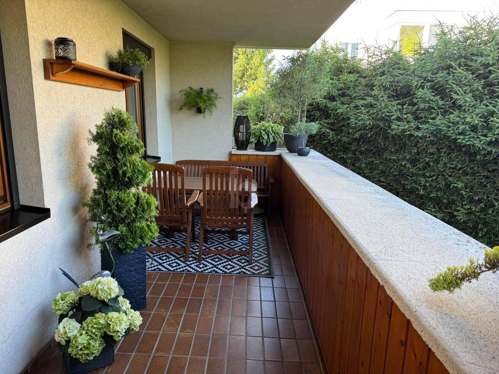 Geräumiger Balkon mit Terrakottafliesen, Holzmöbeln und grüner Hecke.