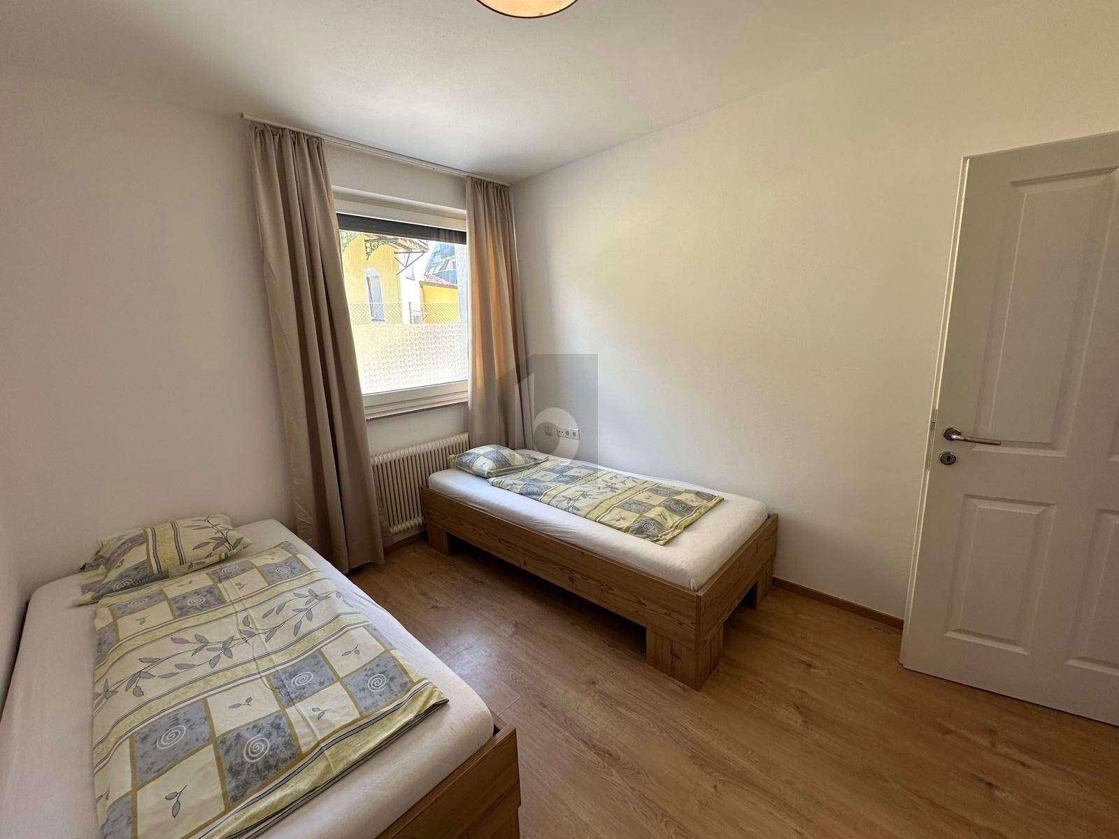 Gemütliches Schlafzimmer mit zwei Holzbetten, hellem Holzboden und Fenster mit Vorhängen.