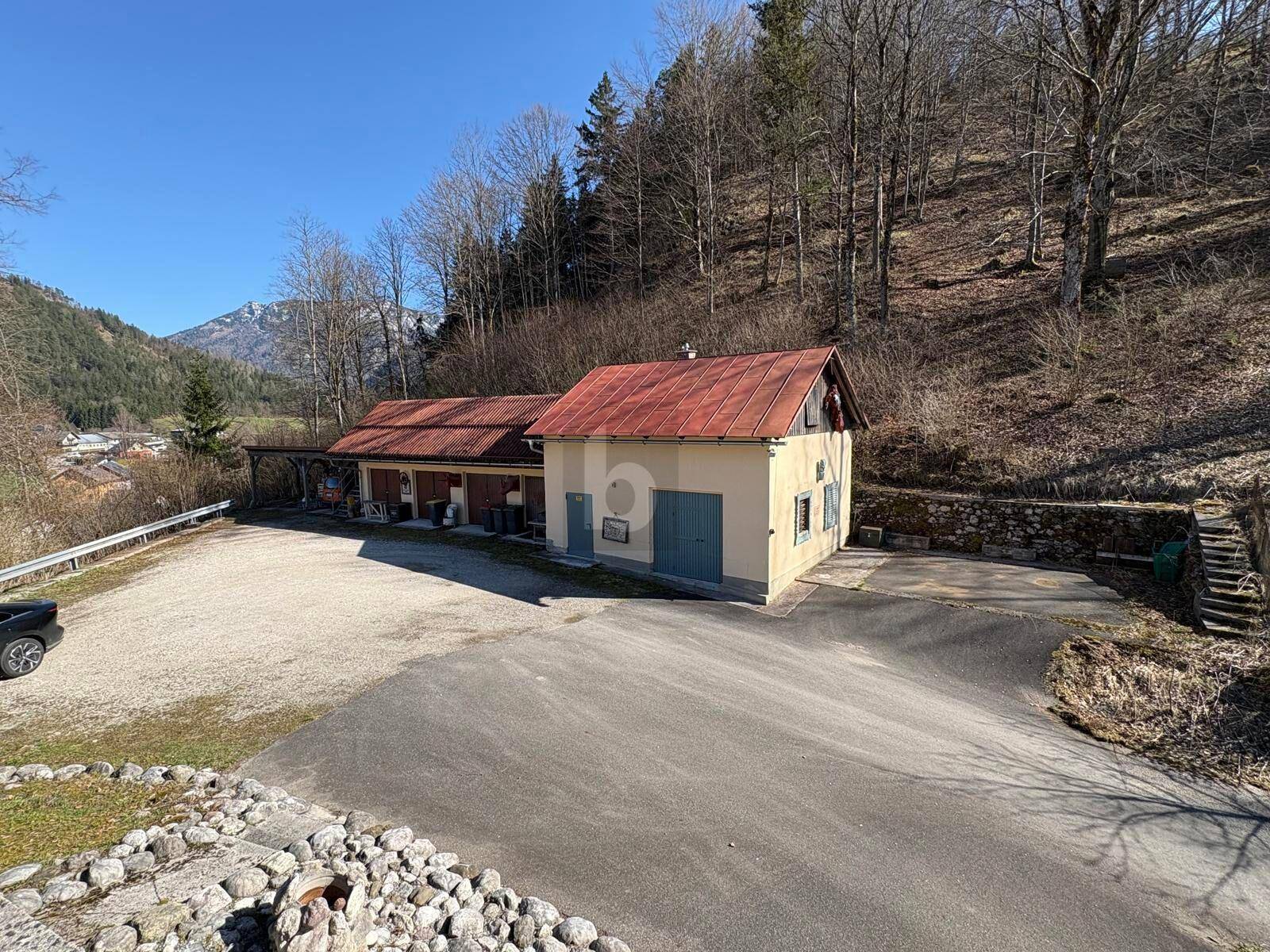 Gebäude mit rotem Dach und großem Parkplatz vor bewaldetem Hang und Bergen im Hintergrund.