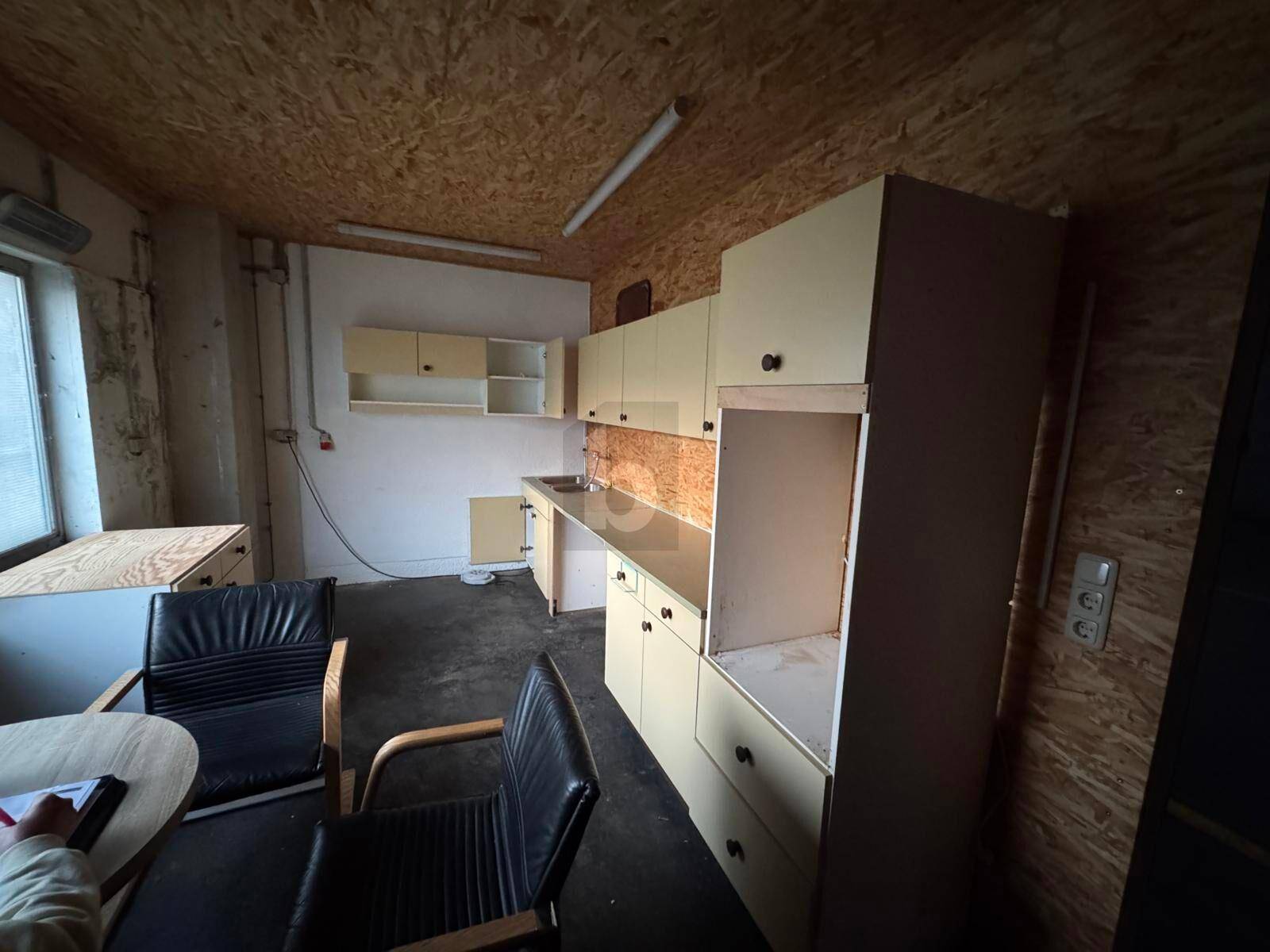 Kleine Küche oder Pausenraum mit älteren Schränken, Spüle und einem Tisch mit Stühlen.