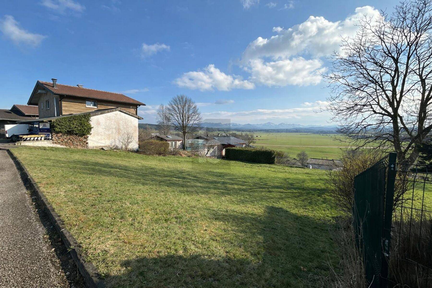 Grüne Wiese mit einem Wohnhaus im Hintergrund und weitem Blick auf die Bergkette.