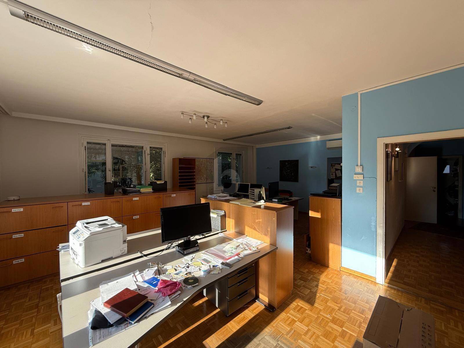 Helles Büro mit Parkettboden, blauer Akzentwand und Zugang zu einem weiteren Raum, gut beleuchtet.