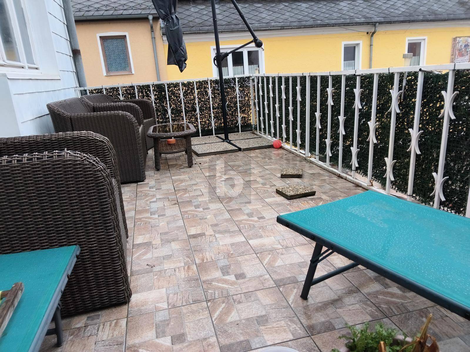Geflieste Terrasse mit Korbmöbeln und einem Sonnenschirm, umgeben von einer Hecke.