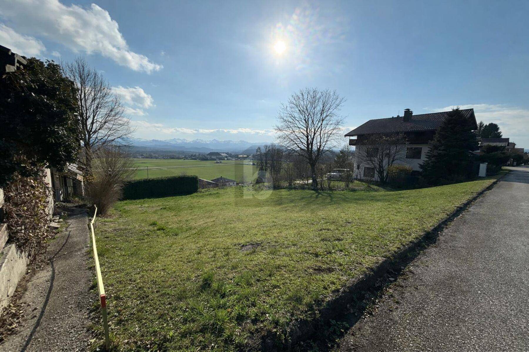Sonnige Grünfläche mit einem Haus am Rand und Panoramablick auf die umliegende Berglandschaft.