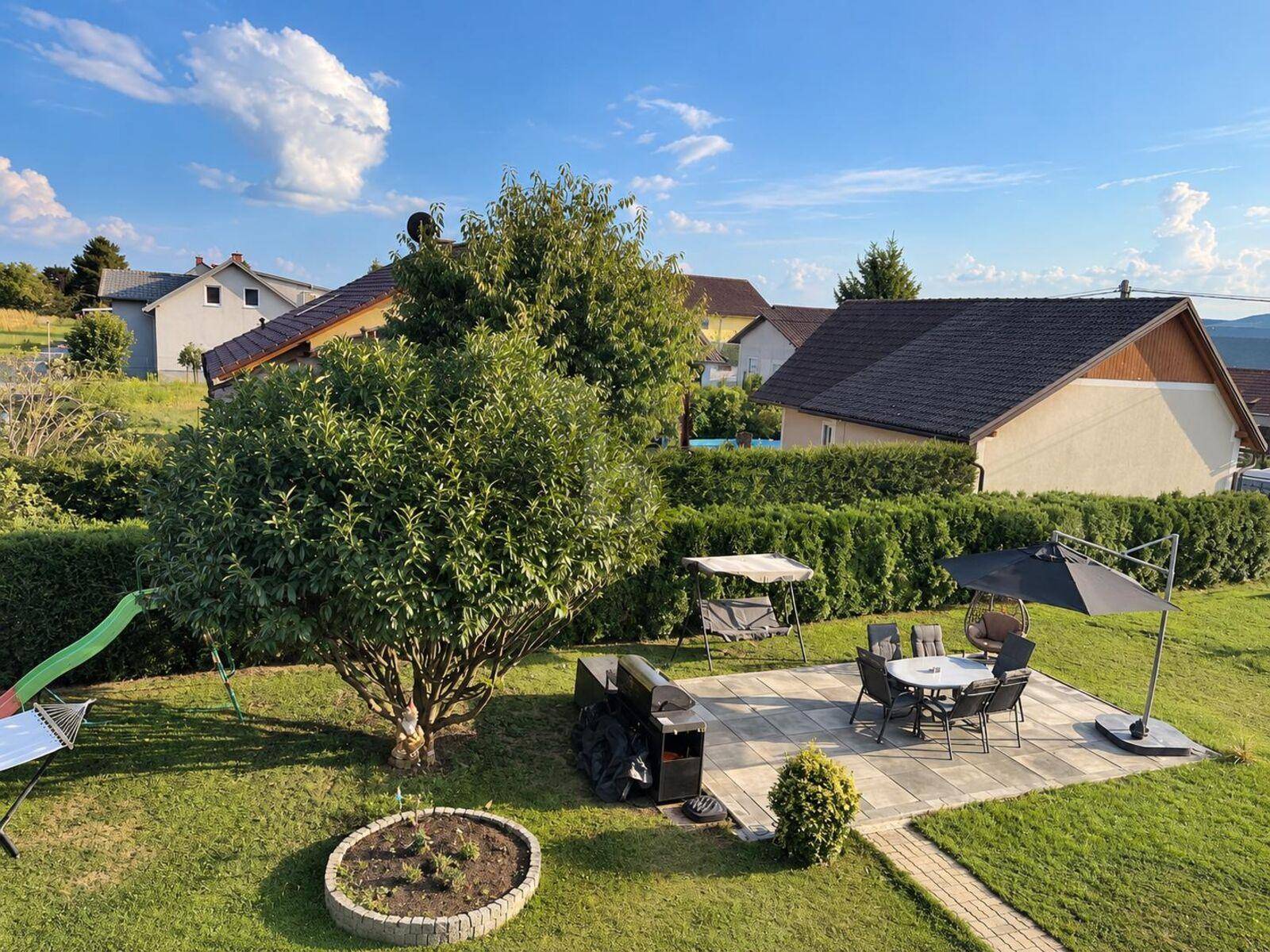 Gepflegter Garten mit Rasenfläche, Sitzbereich, Grill und Kinderspielgeräten unter blauem Himmel.