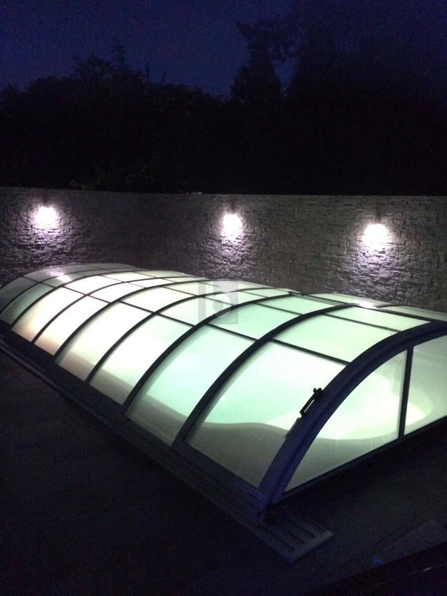 Überdachter Swimmingpool im Außenbereich bei Nacht, beleuchtet von Wandlampen.