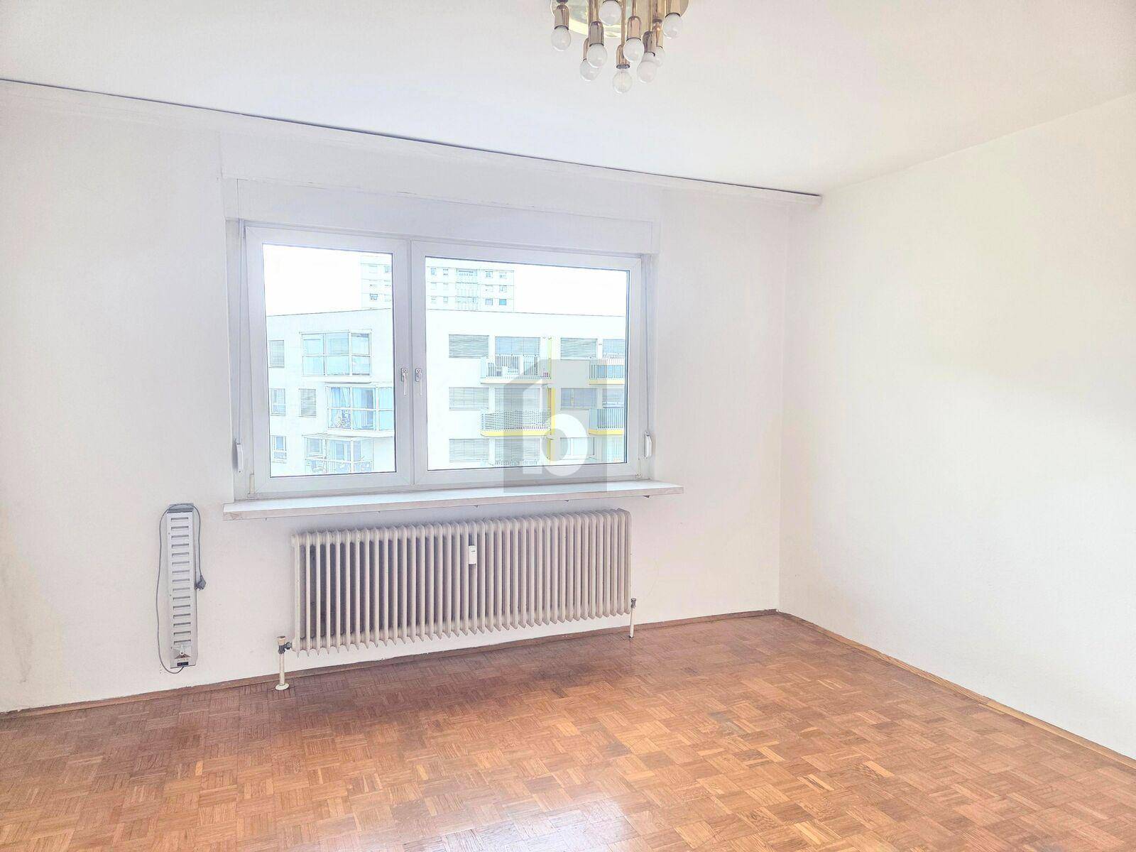Helles Schlafzimmer mit klassischem Parkettboden und großem Fenster mit Blick auf umliegende Gebäude.