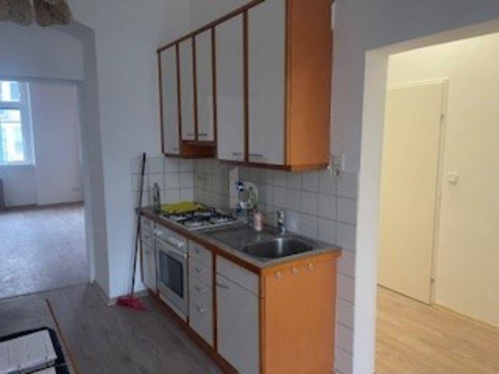 Einfache Küche mit Gasherd, Spüle und orange-weißen Schränken, mit Blick in einen angrenzenden Raum.