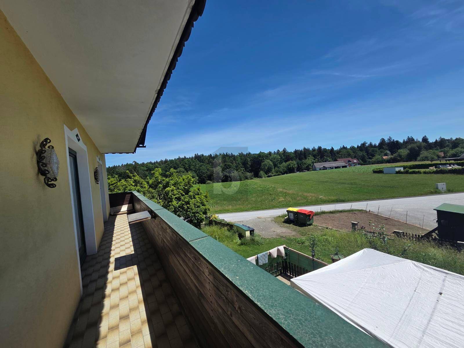 Balkon mit Blick auf eine weite grüne Landschaft und bewaldete Hügel unter strahlend blauem Himmel.