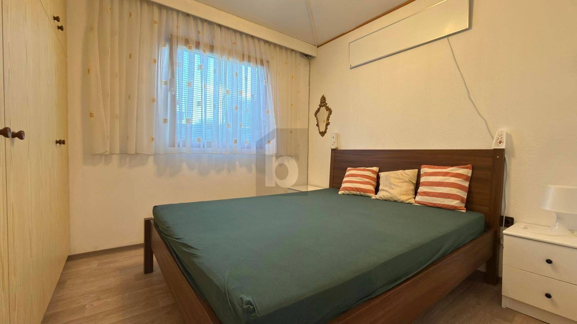 Schlafzimmer mit Holzbett, Einbauschrank und Klimaanlage, hell durch großes Fenster.