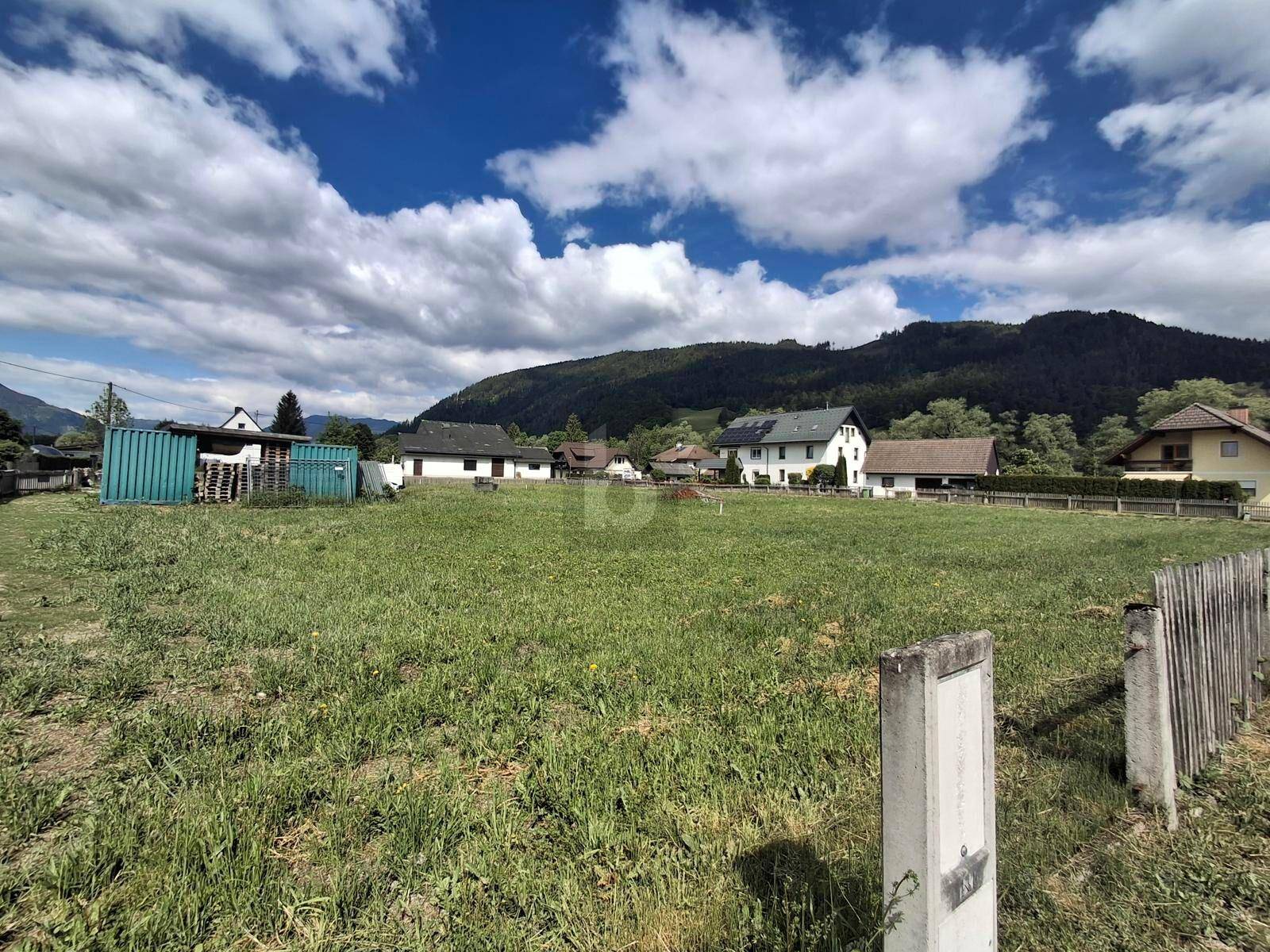 Grünes Grundstück mit umliegenden Häusern und majestätischen Bergen unter einem bewölkten Himmel.