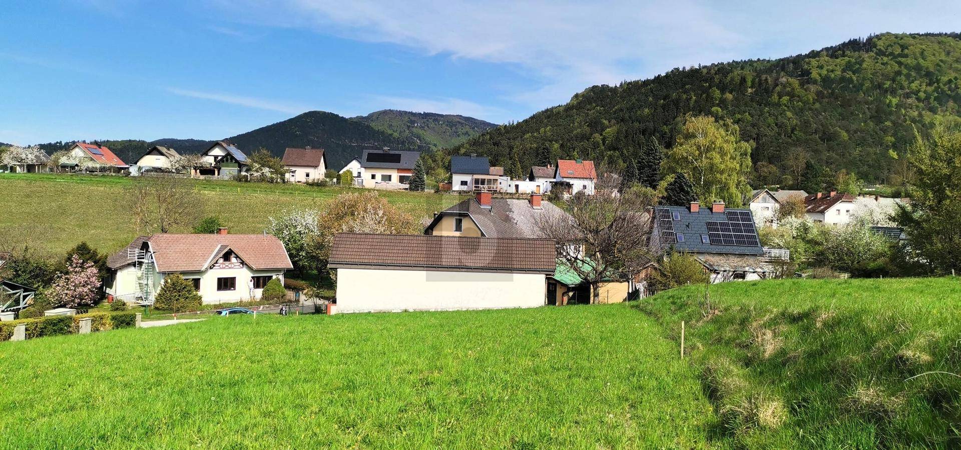 Weitläufige Ansicht einer Wohnsiedlung in ländlicher Umgebung mit grünen Wiesen und bewaldeten Hügeln.