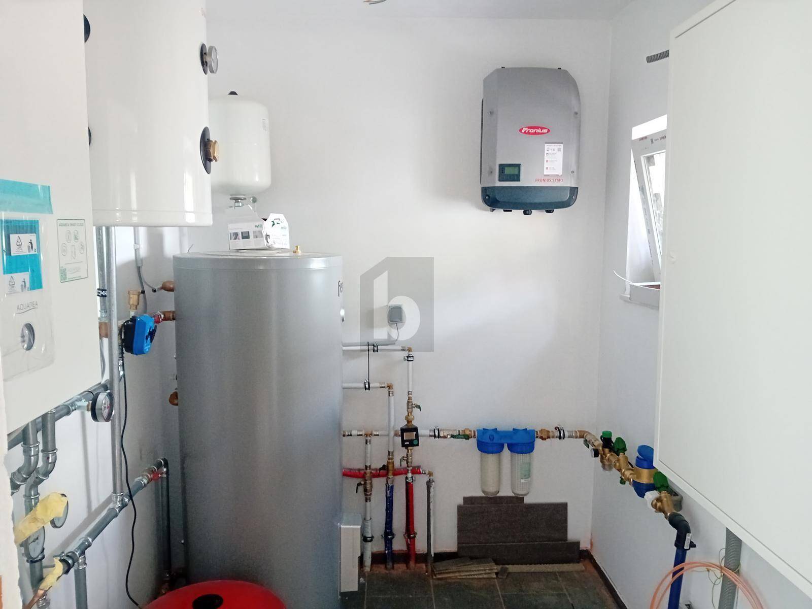 Technikraum mit Warmwasserspeichern, Rohrleitungen und einem Wechselrichter an der Wand.