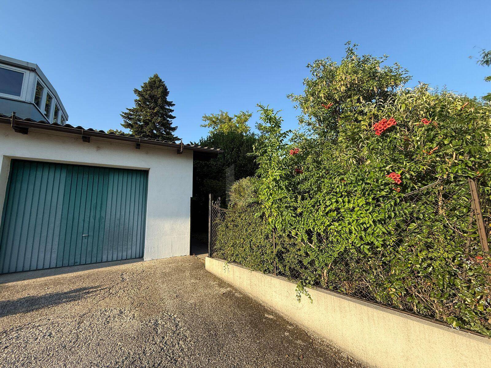 Eine Garage mit grünem Tor und Zufahrt, umgeben von üppigem Grün und blühenden Pflanzen.