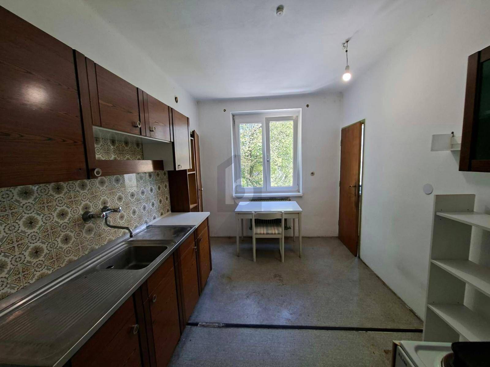 Ältere Küche mit Holzschränken, Spüle und einem kleinen Esstisch am Fenster.