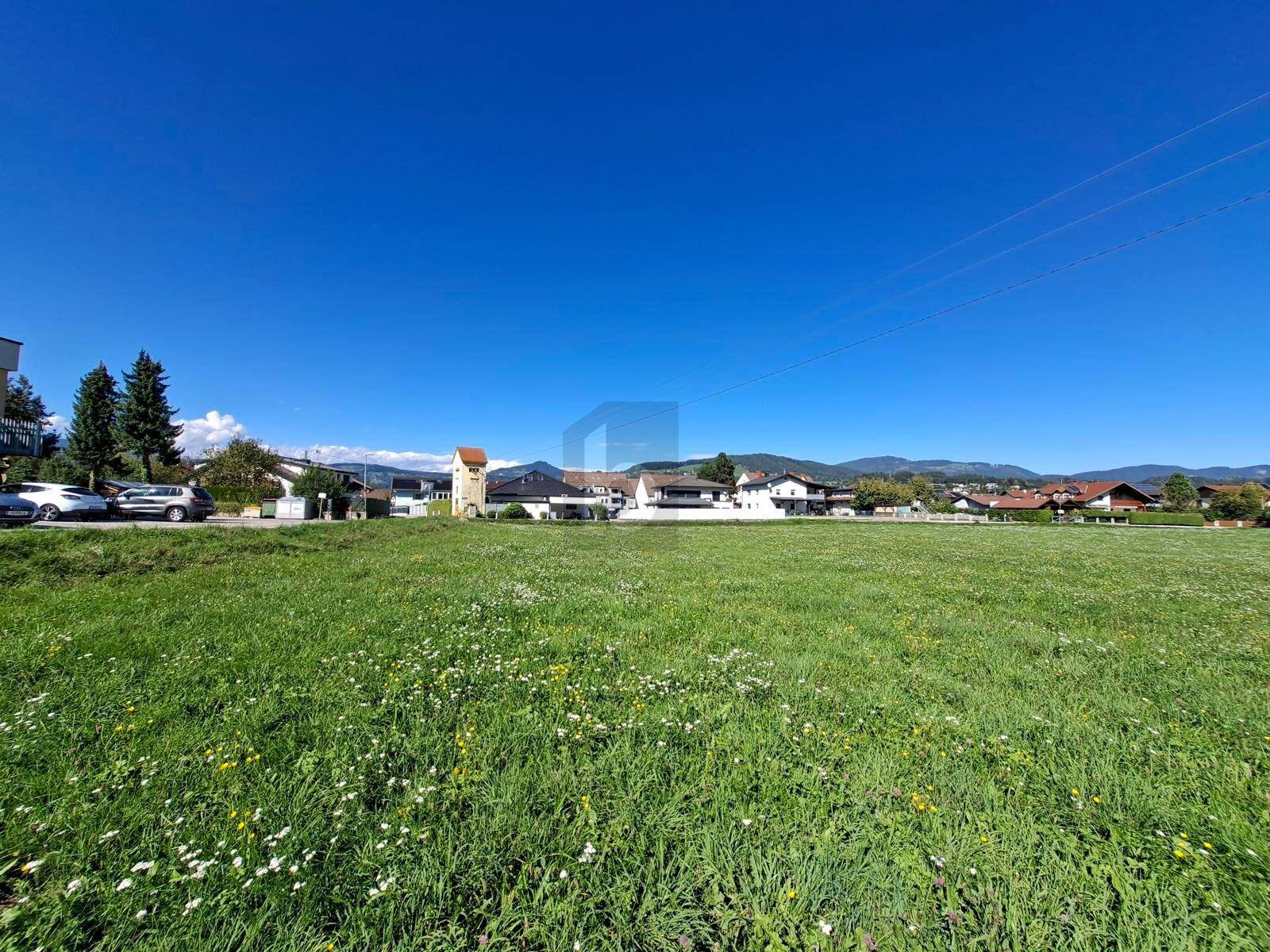 Breites grünes Feld mit Häusern und Bergen in der Ferne, beleuchtet von der Sonne unter blauem Himmel.