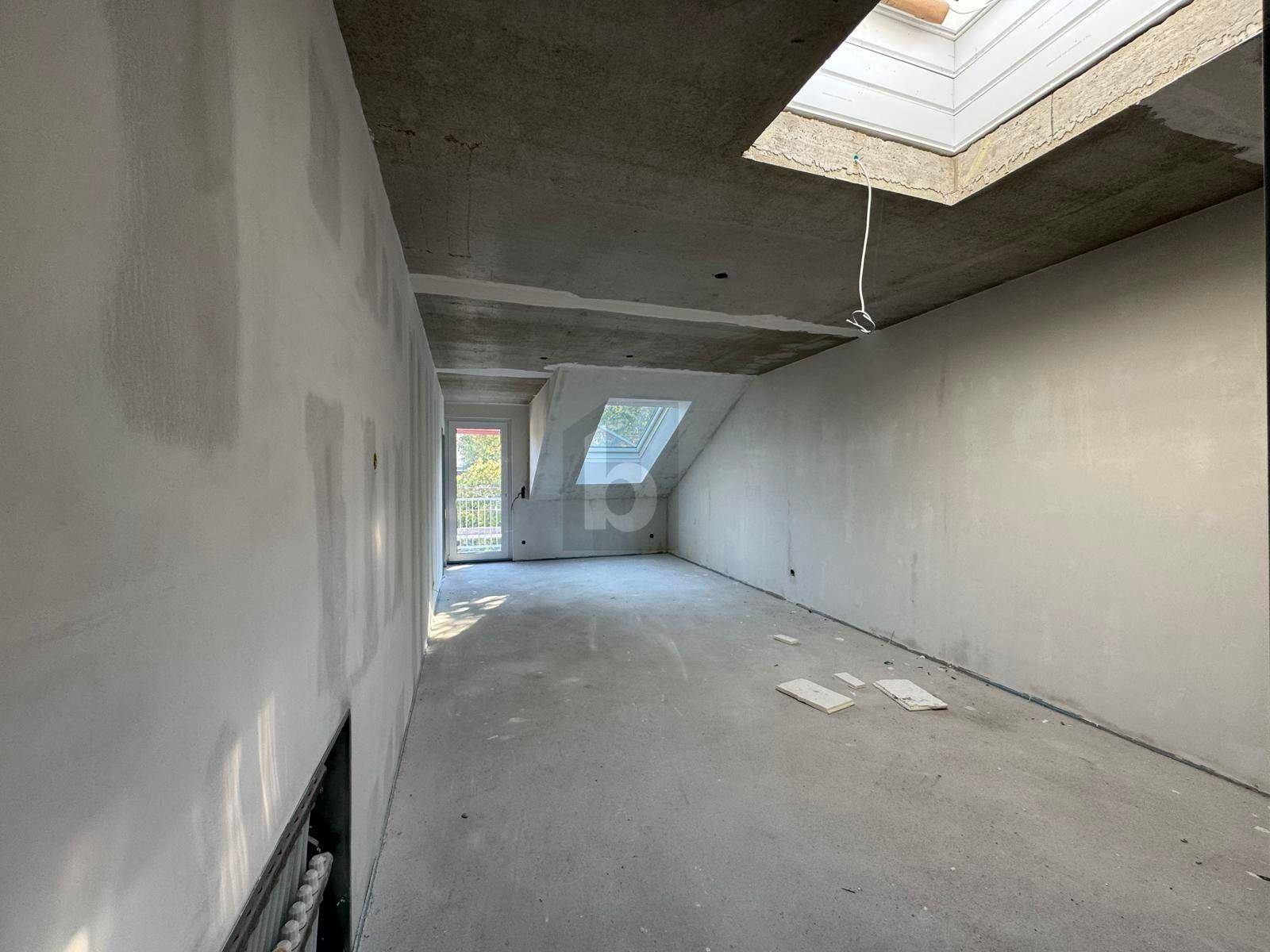 Unfertiger Innenraum im Dachgeschoss mit schrägen Wänden, Dachfenster und Zugang zu einem Balkon.