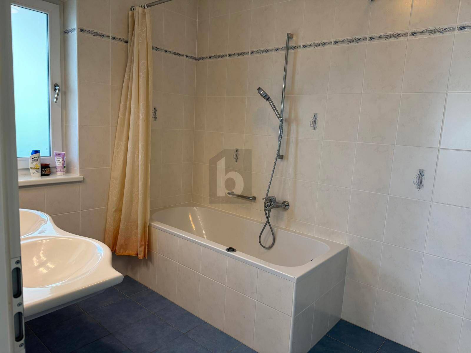Badezimmer mit Badewanne, Waschbecken, hellen Fliesen und Fenster für Tageslicht.