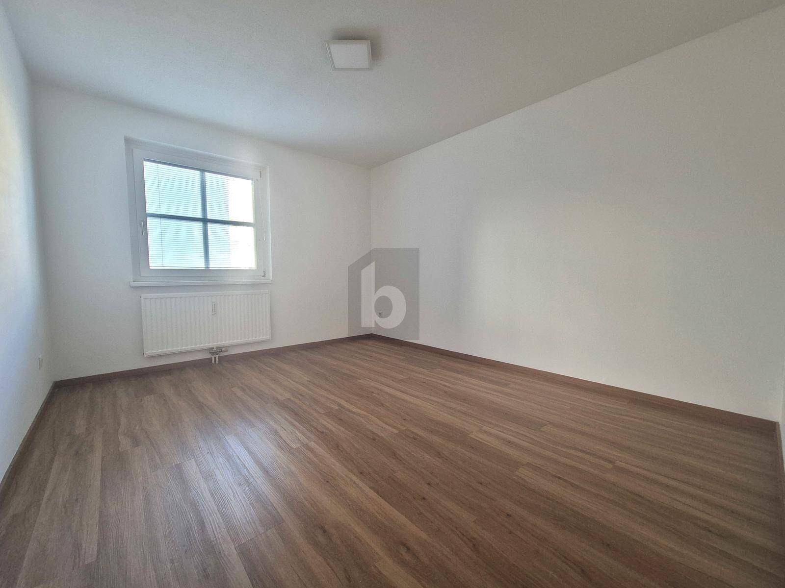 Helles, leeres Zimmer mit Laminatboden und Fenster, bereit zur individuellen Gestaltung.