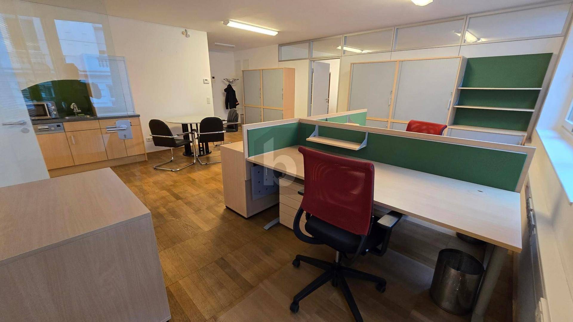Bürofläche mit integrierter Küchenzeile, mehreren Arbeitsplätzen und einem modernen roten Bürostuhl.