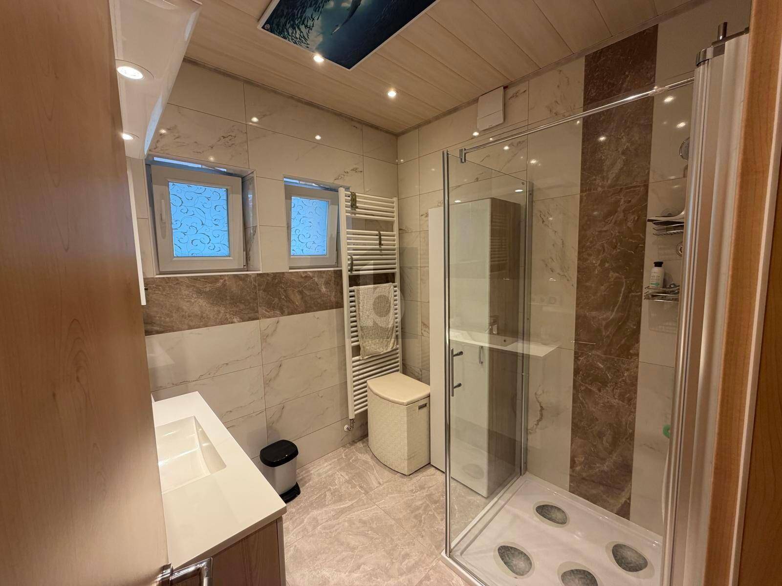 Modernes Badezimmer mit begehbarer Dusche, Einzelwaschbecken, beheiztem Handtuchhalter und zwei Fenstern.