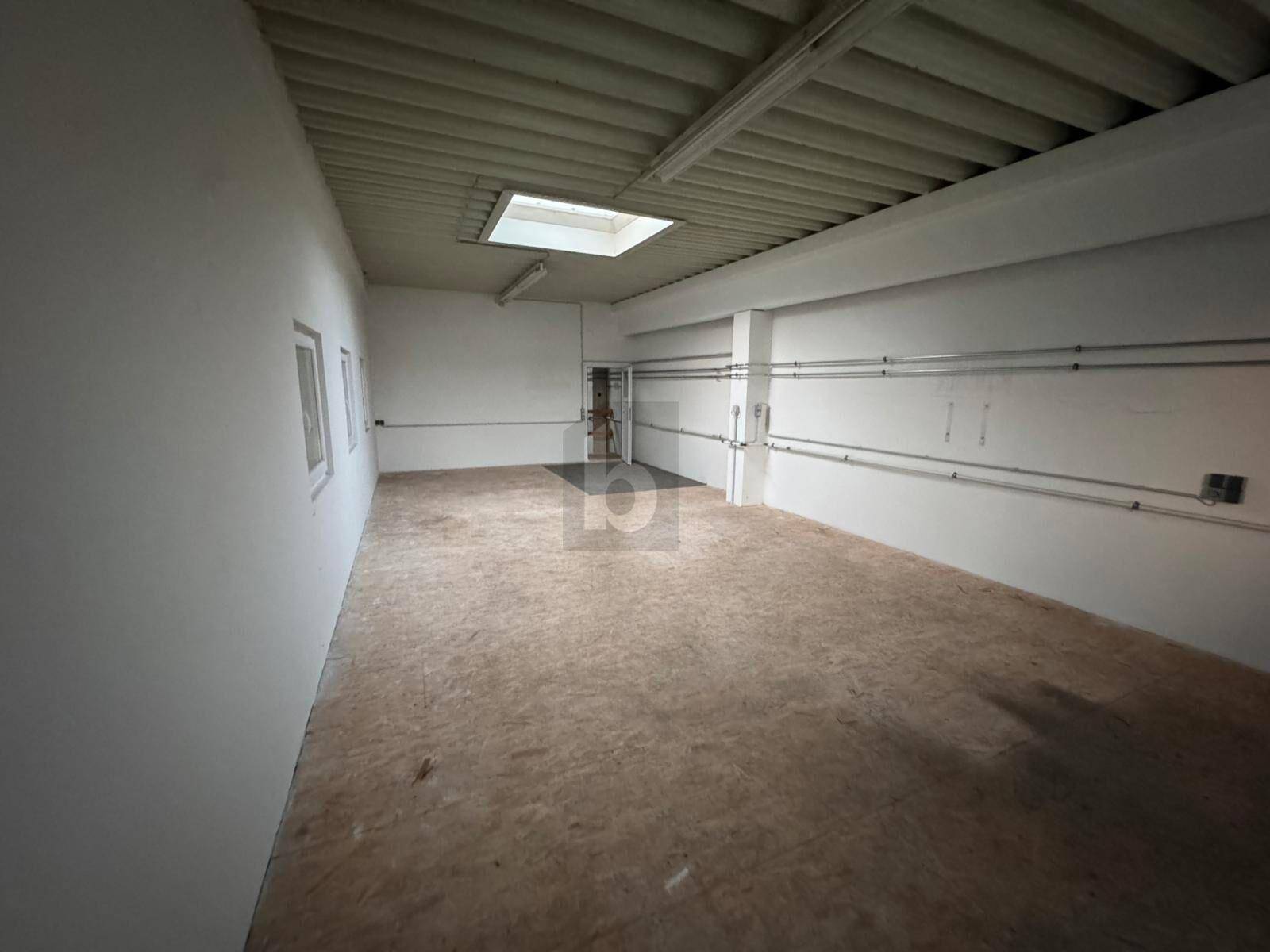 Heller, leerer Raum mit Spanplattenboden, weißen Wänden und sichtbaren Leitungen an der Seite.