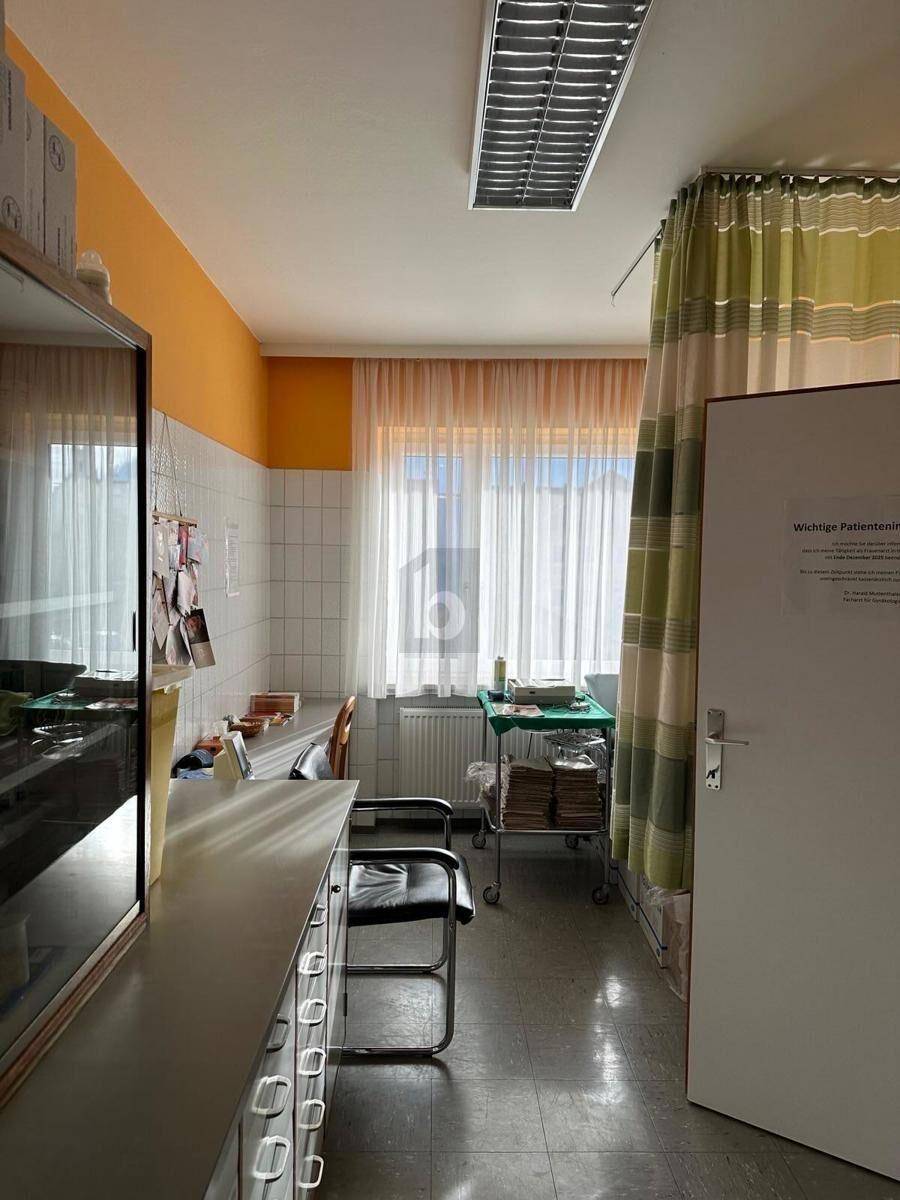 Behandlungsraum mit langer Arbeitsplatte, gefliestem Wandbereich und einem Fenster mit Vorhängen.