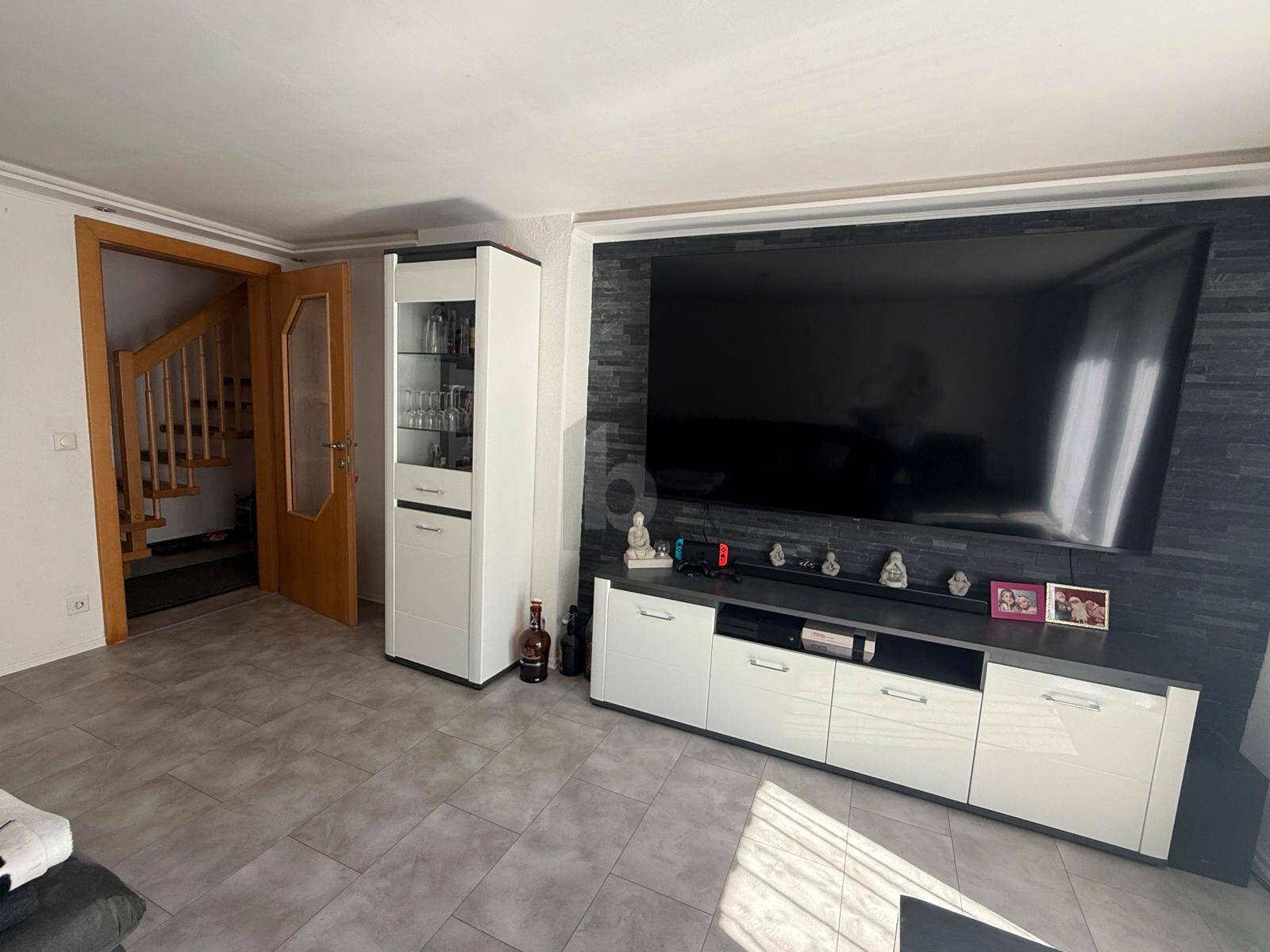 Modernes Wohnzimmer mit großem Fernseher an einer dunklen Steinwand und hellen Fliesenboden.