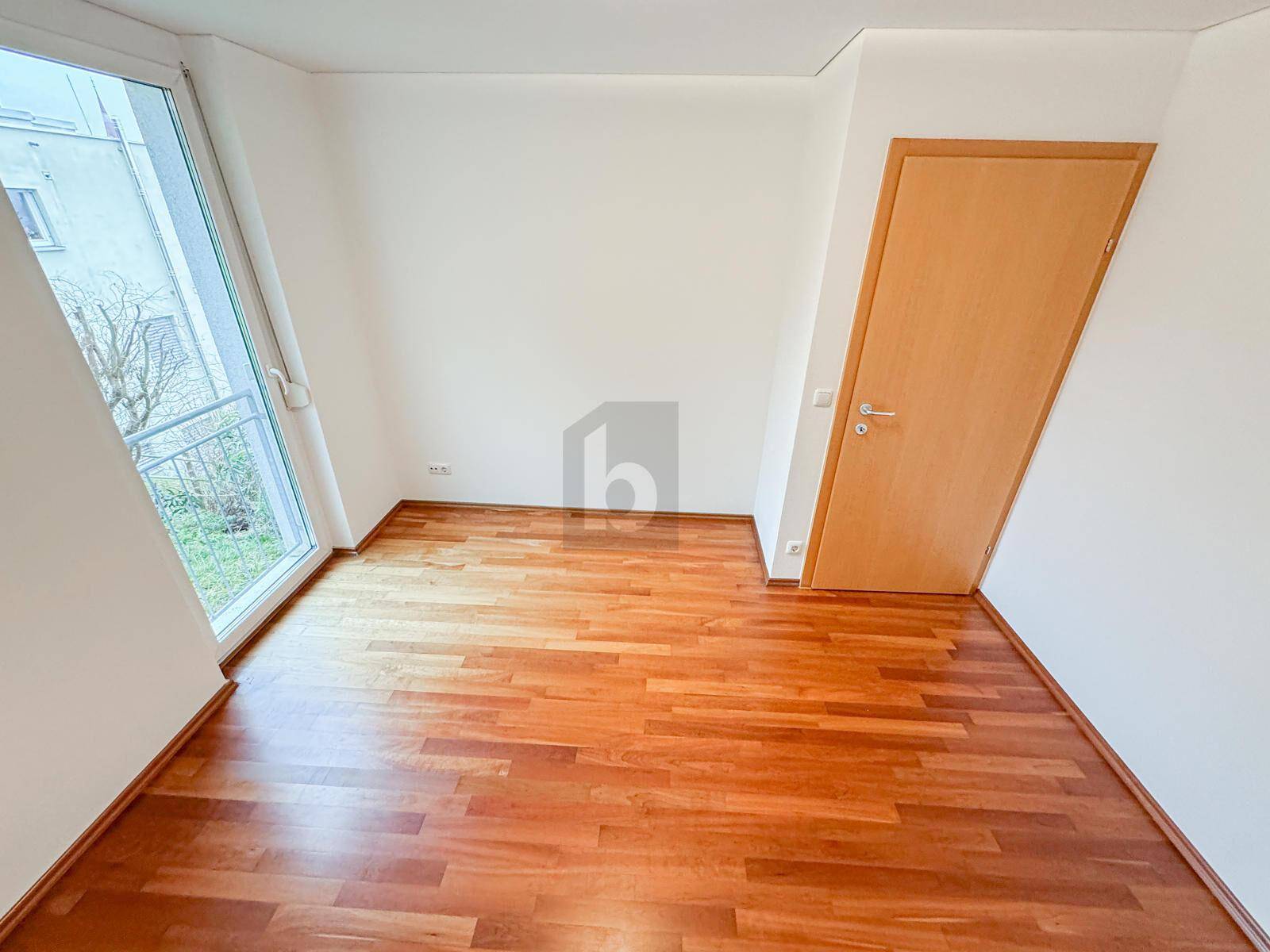 Leeres Zimmer mit Holzboden, weiß gestrichenen Wänden und Zugang zu einem französischen Balkon.