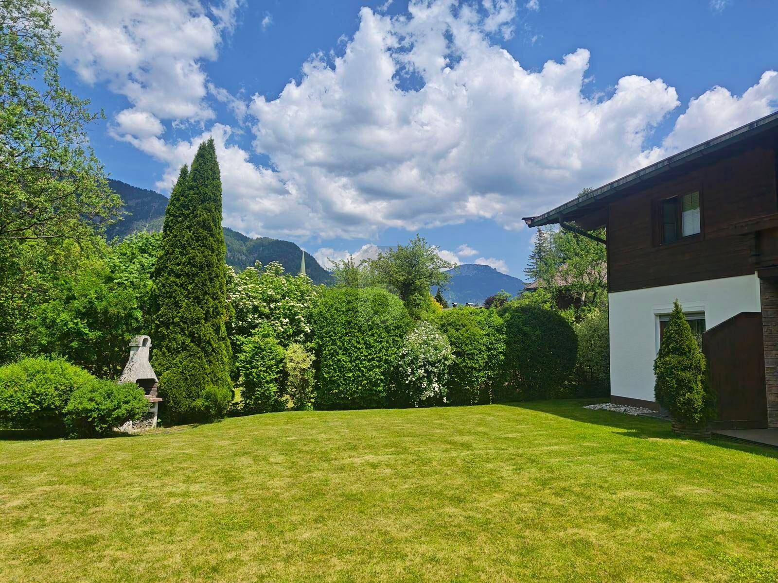 Sonnige Gartenanlage mit großem Rasen, Grillstelle und Blick auf die umliegende Berglandschaft.