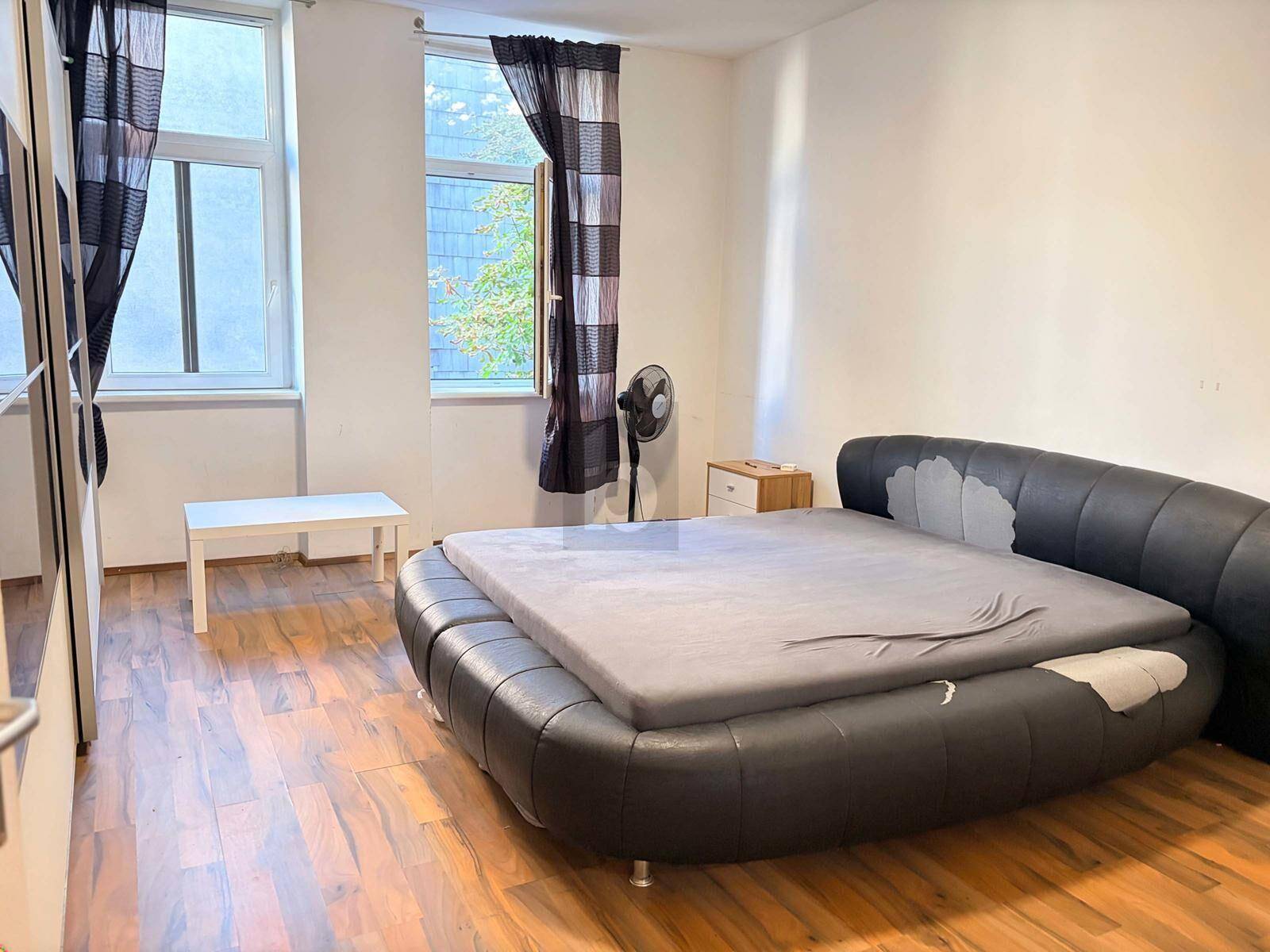 Schlafzimmer mit großem Doppelbett, Laminatboden und zwei Fenstern mit Vorhängen.