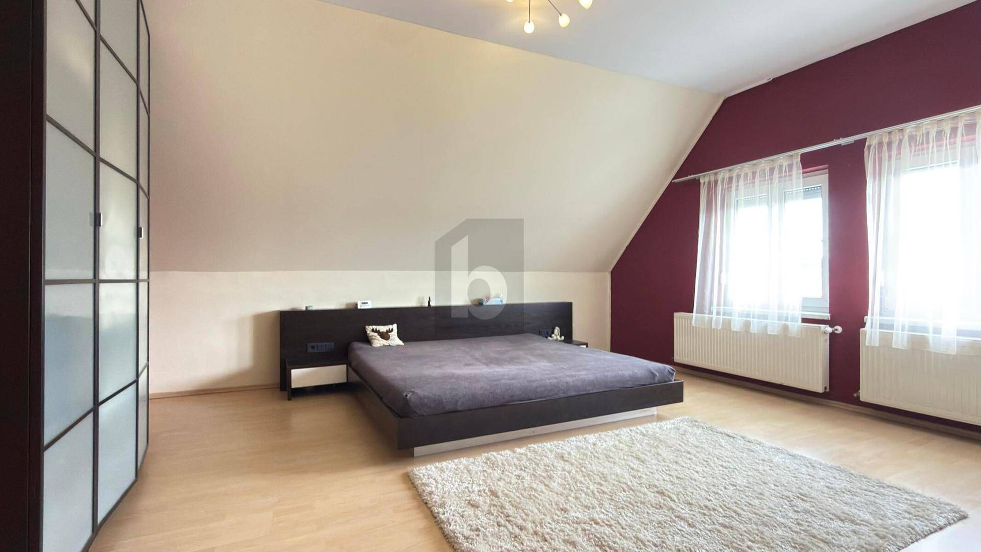 Helles Schlafzimmer mit Dachschräge, großem Bett, Einbauschrank und roter Akzentwand.