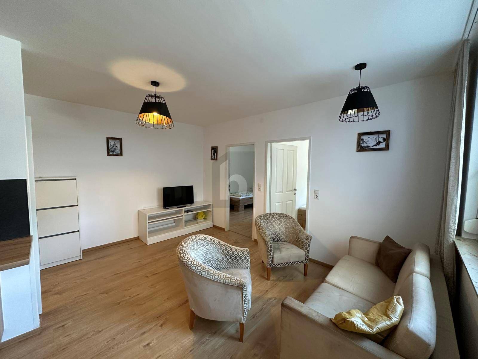 Heller Wohnbereich mit Sofa, zwei Sesseln, TV-Möbel und Zugang zu weiteren Räumen.