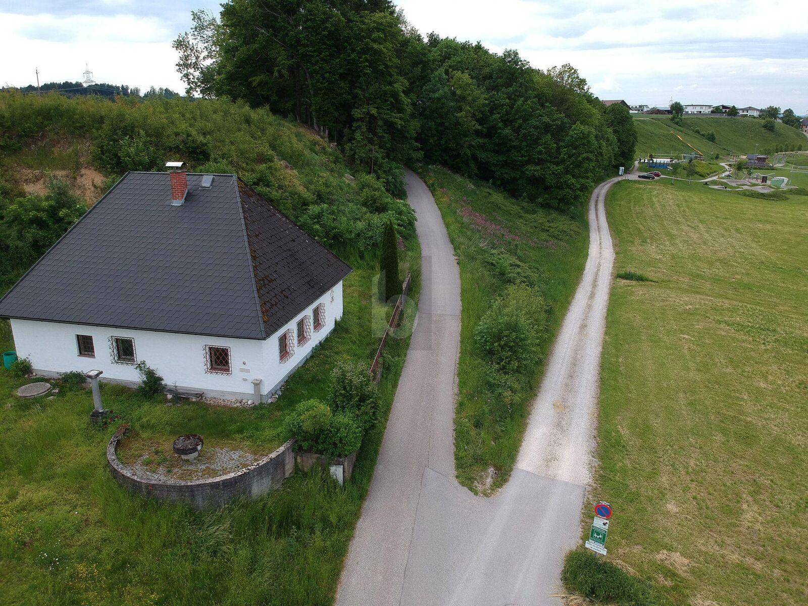 Luftaufnahme eines weißen Hauses mit dunklem Dach, umgeben von grünen Hügeln und zwei Feldwegen.
