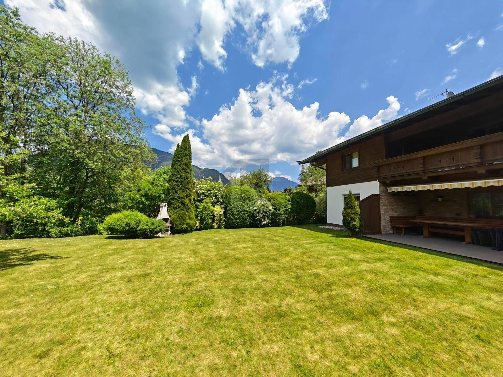 Weitläufiger Garten mit gepflegtem Rasen, Grillplatz und überdachtem Sitzbereich am Haus mit Bergblick.