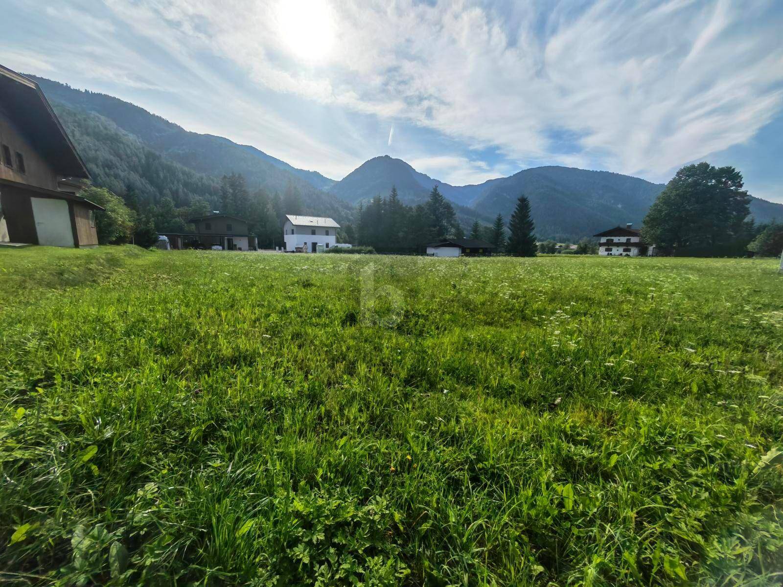 Sonnige Wiesenfläche mit Häusern im Hintergrund und einer beeindruckenden Bergkulisse.