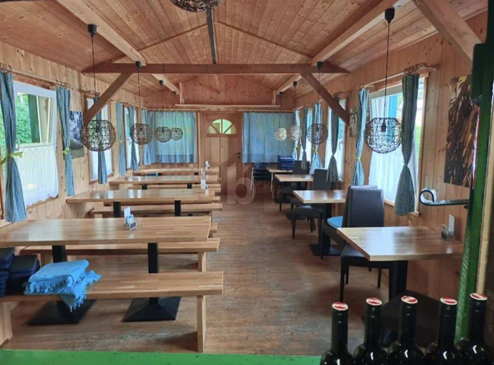 Rustikaler Speisesaal mit Holzwänden, Holzbänken und Tischen, beleuchtet von modernen Hängelampen.