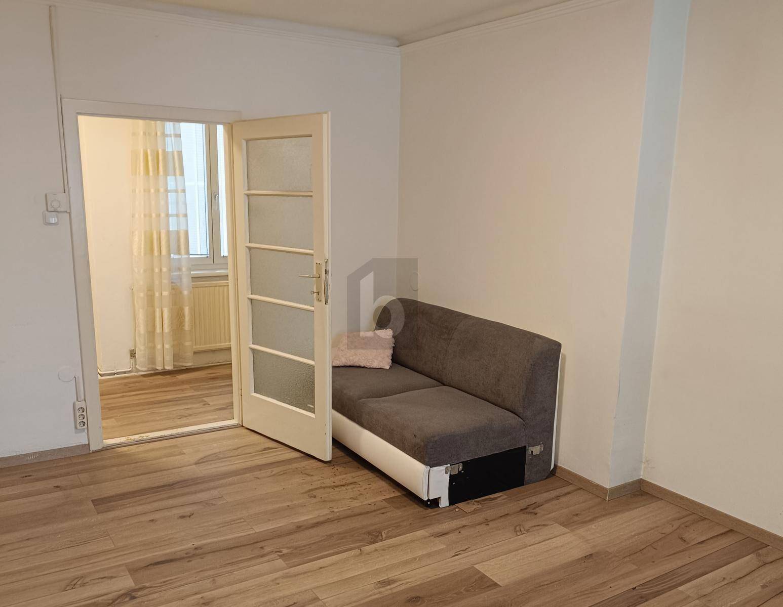 Wohnraum mit Holzoptikboden, weißer Couch und offener Tür zu einem weiteren Zimmer.