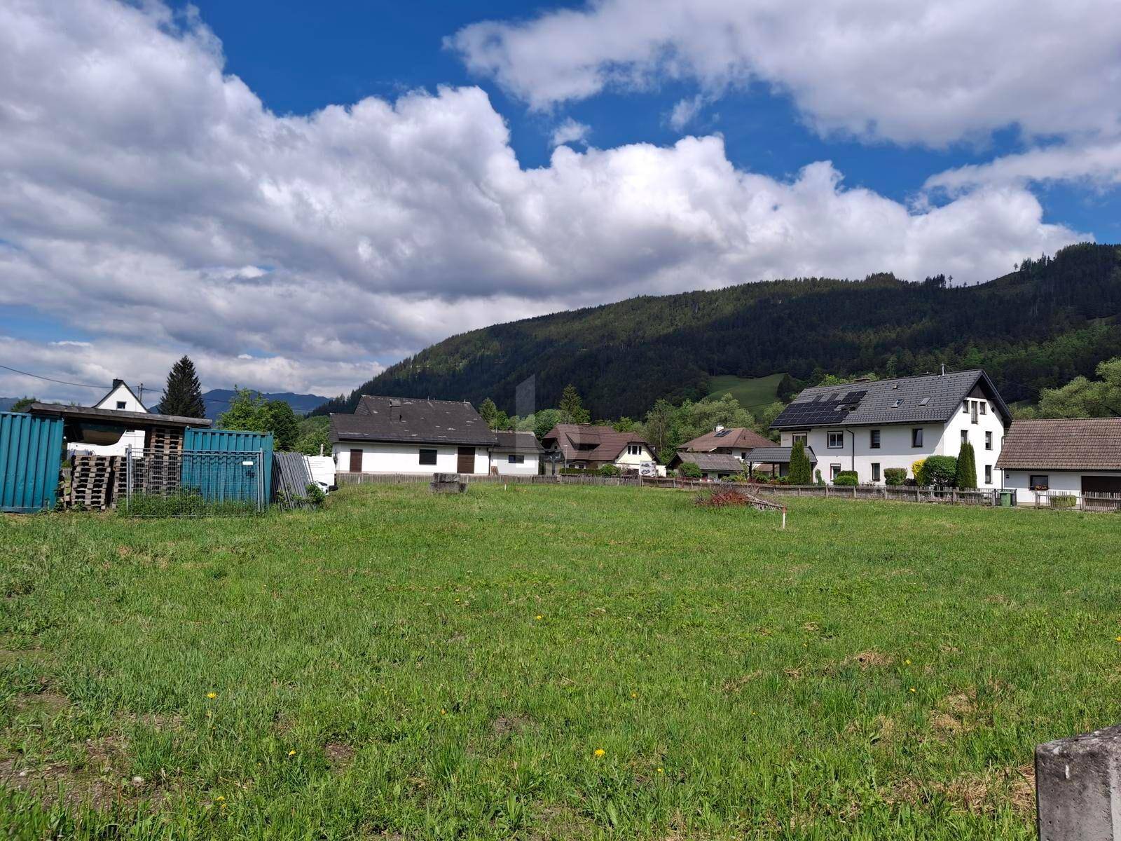 Sonnige Wiese mit Blick auf umliegende Häuser und bewaldete Berge unter einem blauen Himmel.