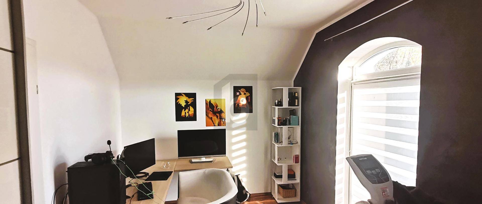 Helles Arbeitszimmer im Dachgeschoss mit Schreibtisch, Computern und einem Fenster mit Rundbogen.
