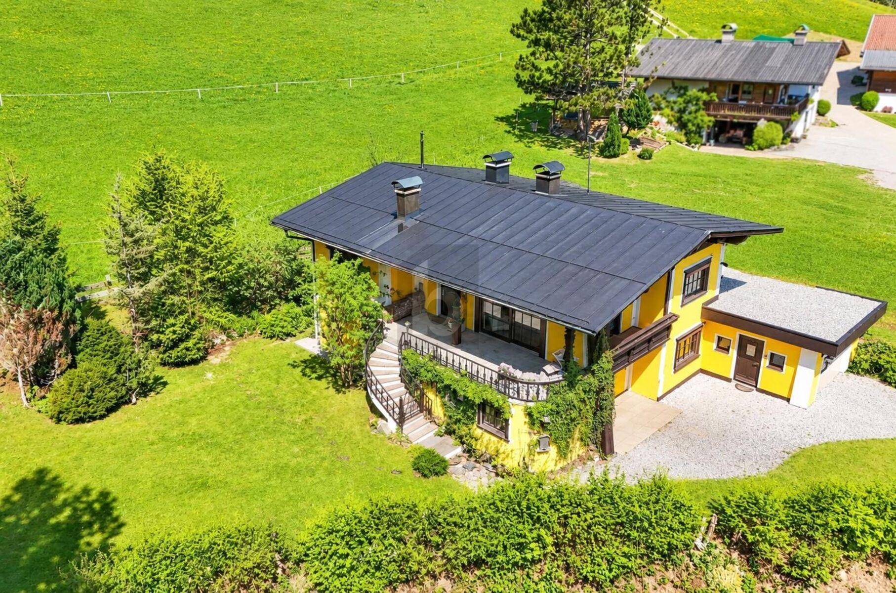 Luftaufnahme eines gelben Hauses mit schwarzem Dach, umgeben von grünen Wiesen und Bäumen. Eine geschwungene Treppe führt zum Eingang.