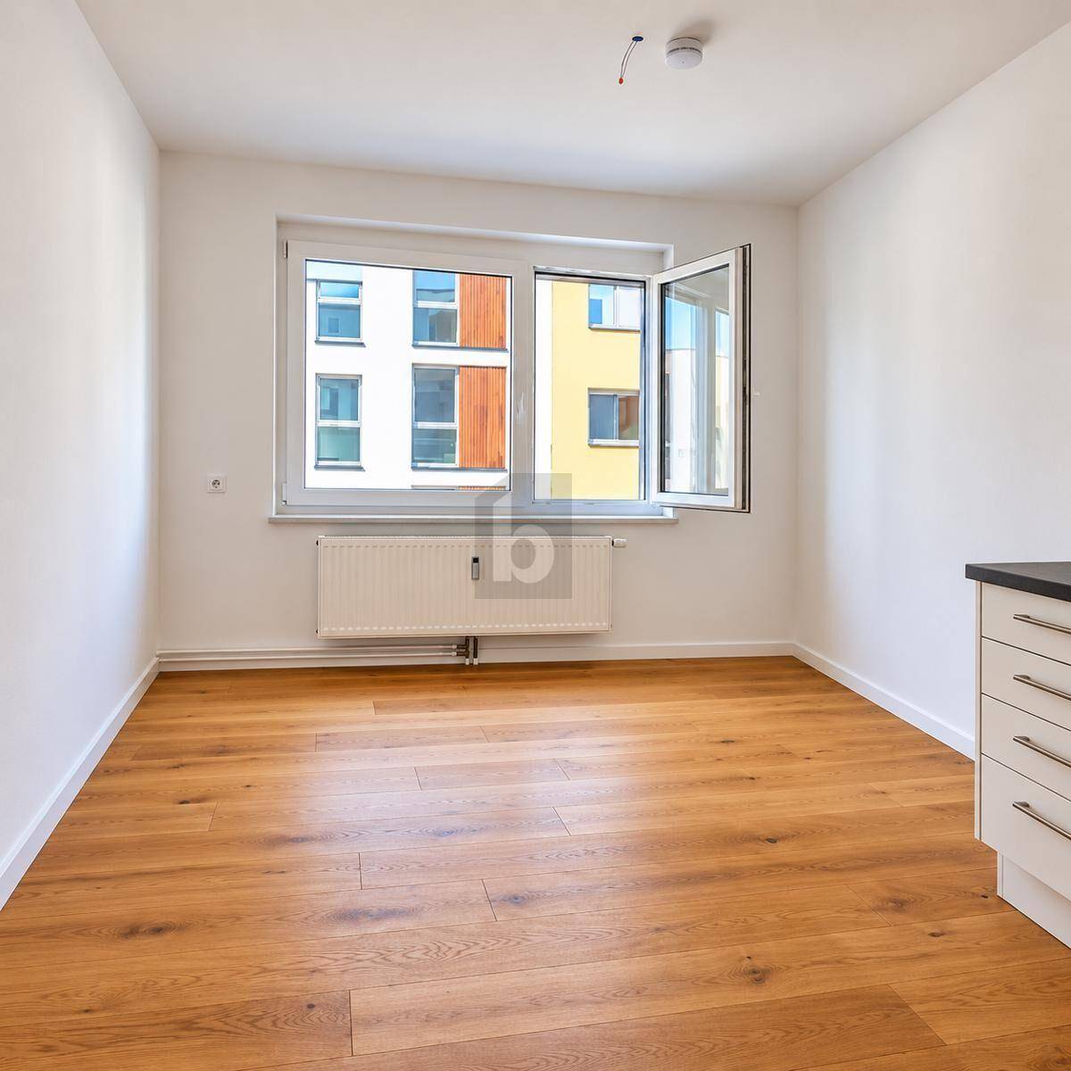 Heller, leerer Raum mit Holzboden, Heizkörper unter dem Fenster und Blick auf andere Gebäude.